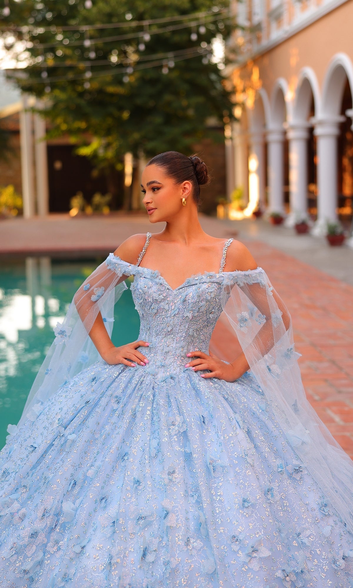 A young woman stands by a pool, wearing the Amarra Quinceanera Dress 54308, an off-the-shoulder light blue ball gown featuring sheer cape sleeves and elegant 3D floral details.